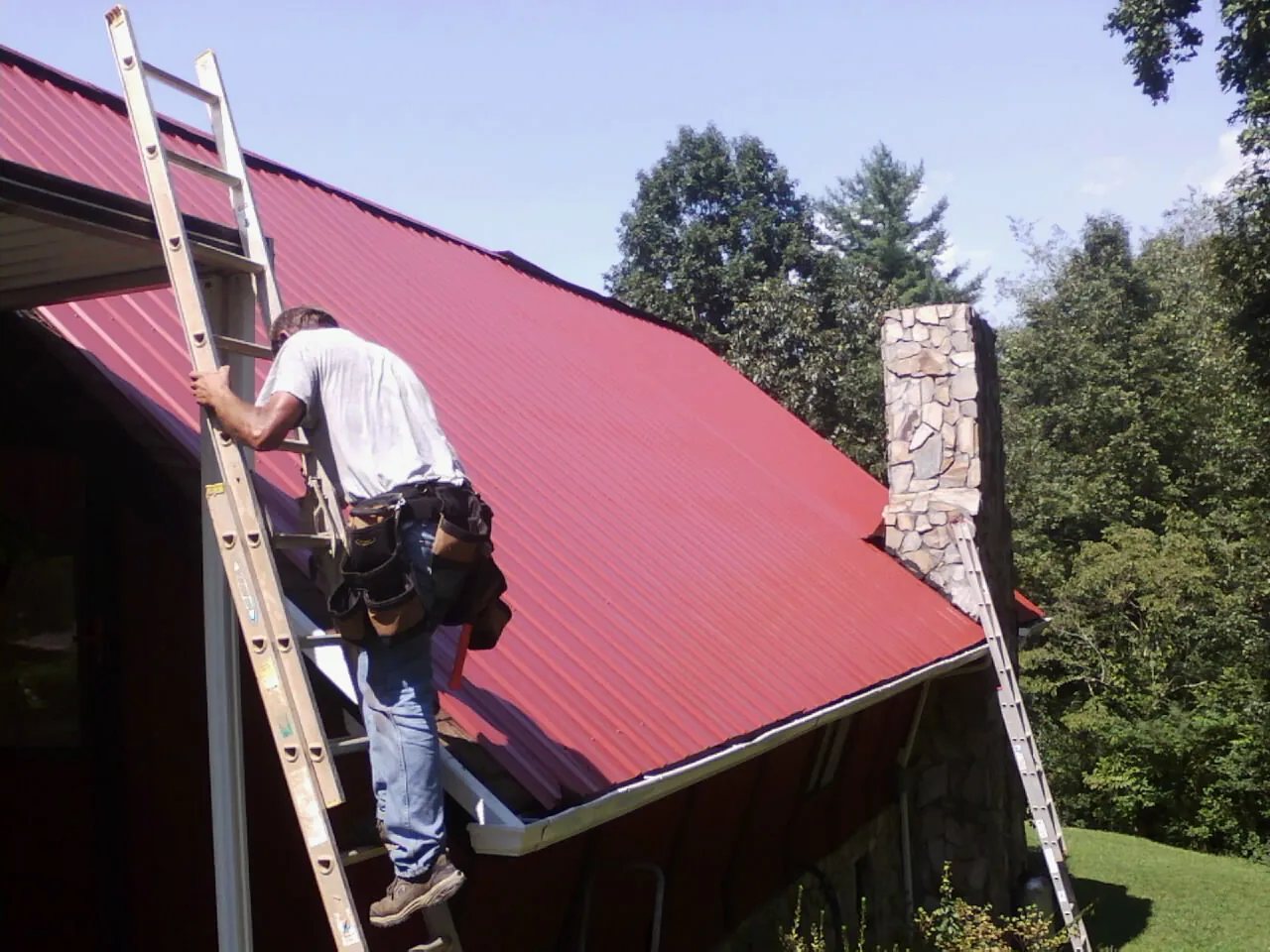 Roofer on a ladder beside a red metal roof and stone chimney — Soffit & Fascia Repair by Roofing Brothers in Genoa